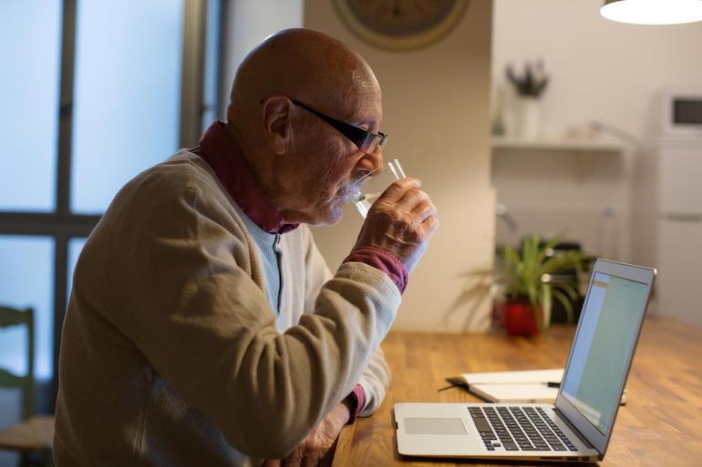 Schonvermögen – was der Pflegebedürftige behalten darf Alter Mann informiert sich über Schonvermögen am Laptop und trinkt ein Glas Wasser